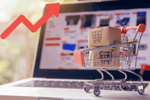 Carrito de compras frente a una computadora que muestra un gráfico de barras en ascenso, simbolizando el aumento en la recaudación por impuestos a compras en línea e importaciones.