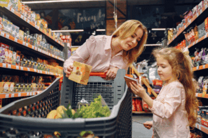 Una mujer y su hija observan un producto en un anaquel de supermercado mientras empujan un carrito de compras.