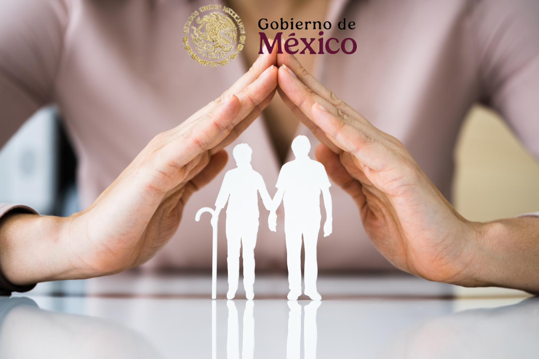 Manos simbólicas cubren a una pareja de ancianos de cartón, representando seguridad y protección, con el logotipo del Gobierno de México al fondo.